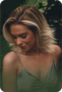 A beautiful woman with blonde hair and glowing skin, wearing a gray spaghetti strap top, looks downward against a backdrop of lush greenery. - Bridge Aveda Salon | Chattanooga, TN